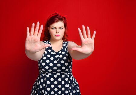 Serious young caucasian size woman plus showing stop gesture with her hand. She dressed in a polka dot dress in the studio on a red backgroundの写真素材