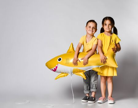 Positive little children, dressed in festive clothes, wears a balloon in the shape of a yellow shark fish, celebrates a holiday, has a wide smile, stands on a light background, being in a good mood. Children and holiday conceptの写真素材