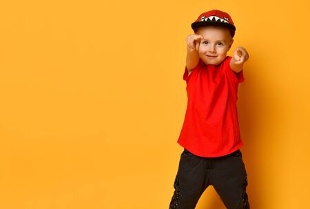 Hipster toddler boy in a red T-shirt and dark sweatpants white sneakers and a funny cap pointing to you on a yellow backgroundの写真素材