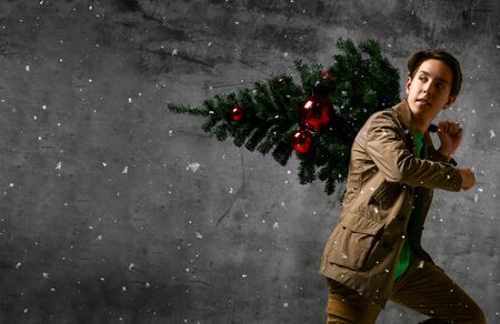 Teenager man carrying a Christmas tree. Young man in a jacket carrying a Christmas tree decorated with red Christmas balls. Merry guy holds a Christmas tree on his shoulders. Snowing timeの写真素材