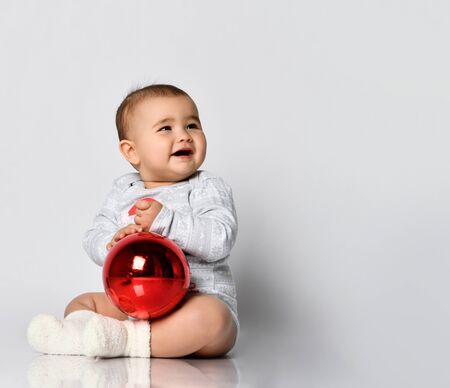 Cute baby girl in grey bodysuit and terry socks plays on the floor with Christmas red balls