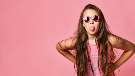 Portrait of young plus size girl with long hair in stylish summer casual clothing and round sunglasses showing tongue and feeling joyful over pink background. Trendy summer youth clothing conceptの写真素材