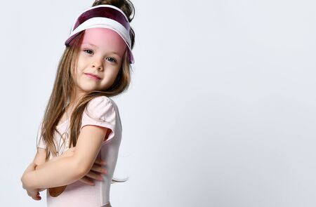 Small smiling cute girl in casual summer dress, cap with a visor standing, looking up and posing over white wall background in photo studio.の写真素材