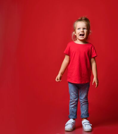 Small happy excited blond girl in stylish casual clothing and white sneakers standing and smiling over red wall background. Trendy children clothing and happy childhood conceptの写真素材