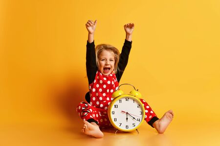 Little happy blond girl in red dotted pajamas sitting on floor with big alarm clock and feeling excited over yellow background. Different times of day and children schedule conceptの写真素材