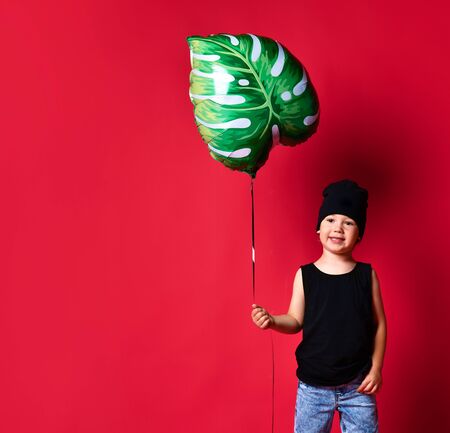 Small positive boy in stylish casual clothing and cap standing and holding leaf printed air balloon in hand over red wall background.の写真素材