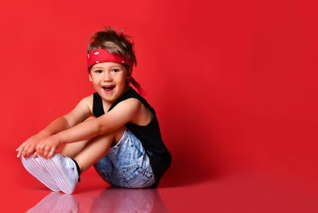 Small happy boy in stylish casual clothing, red hair bandana and white sneakers sitting on floor and smiling over red background.の写真素材