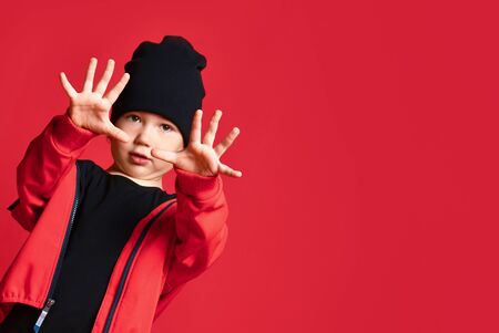 Small positive boy in red jacket and cap standing and showing all his fingers over red wall...