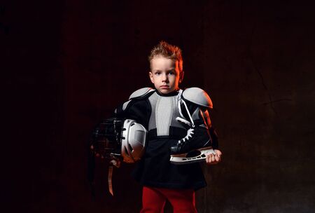 Happy blond boy in hockey uniform with hockey skates and helmet standing and smiling over dark background. Children in sport and young hockey players conceptの写真素材