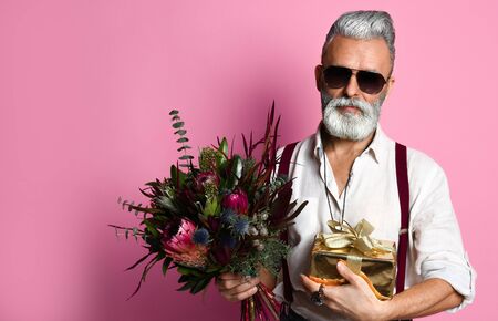 Stylish middle-aged bearded man with a modern haircut, sunglasses and fashionably dressed holds a bouquet of flowers and a gift. Going on a date.の写真素材