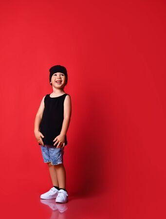 Small positive boy in stylish casual clothing, cap and white sneakers standing and smiling over red wall background. Trendy children clothing and happy childhood conceptの写真素材