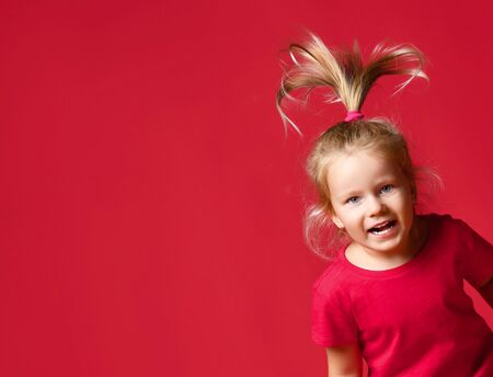 Portrait of jumping small happy excited blond girl in stylish casual clothing with hair raised up over red wall background. Trendy children clothing and happy childhood conceptの写真素材