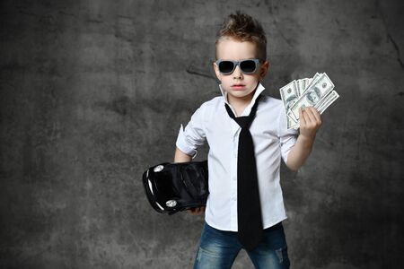 Serious boy little businessman in white shirt, jeans, tie and sunglasses standing and holding black toy car present and dollars cash over grey background. Trendy children clothes, holiday gift conceptの写真素材