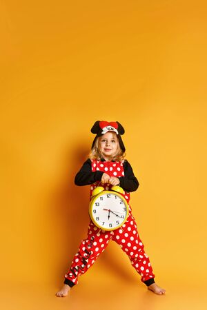 Little happy blond girl in red dotted pajamas standing, holding big alarm clock and feeling excited over yellow background. Different times of day and children schedule conceptの写真素材