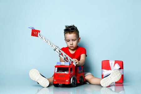 Positive boy in bright stylish casual clothing and sneakers sitting on floor with toy fire engine and feeling happy over blue background. Trendy children clothes, holiday gift, party conceptの写真素材