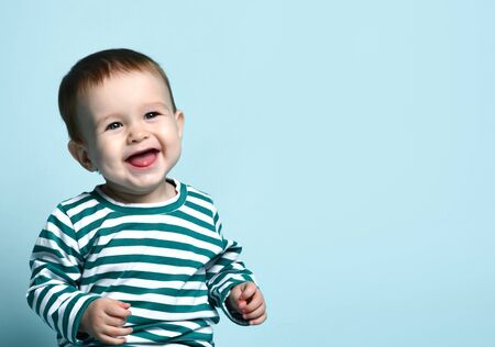 Little baby boy in stylish casual clothing barefoot sitting on floor and smiling over blue wall background. Trendy baby clothing and happy childhood conceptの写真素材