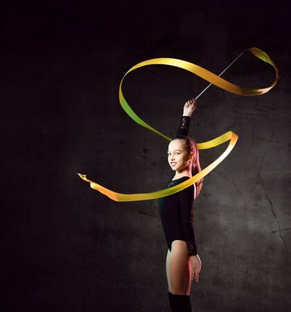 Cute girl gymnast in black sport body and uppers standing and making exercise with yellow gymnastic tape over dark background. Rhytmhic gimnastics beauty cocneptの写真素材