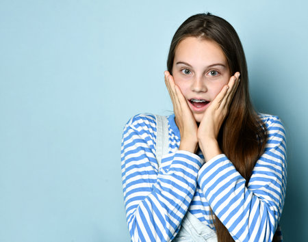 Teen lady in jeans overall, striped sweatshirt. She is looking surprised while posing on blue studio background. Braces on teeth. Sincere emotions, hipster style, fashion, beauty. Copy space. Close upの写真素材
