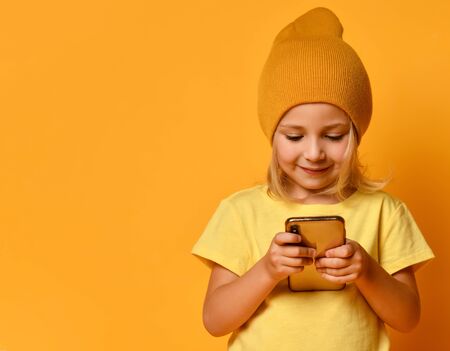 Little blonde kid dressed in t-shirt and hat, holding smartphone, watching videos, looking at screen with joyful smile, posing on yellow studio background. Technology, children, internet. Close-upの写真素材