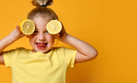 Little smiling cute blond girl in yellow t-shirt holding halves of fresh sour lemon fruit near eyes and showing tongue over yellow background. Healthy lifestyle and clean eating conceptの写真素材