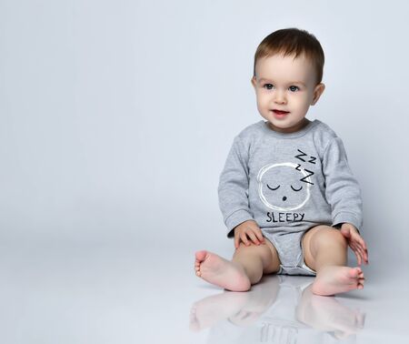 Little baby boy toddler in grey casual jumpsuit and barefoot sitting on floor and smiling over white...