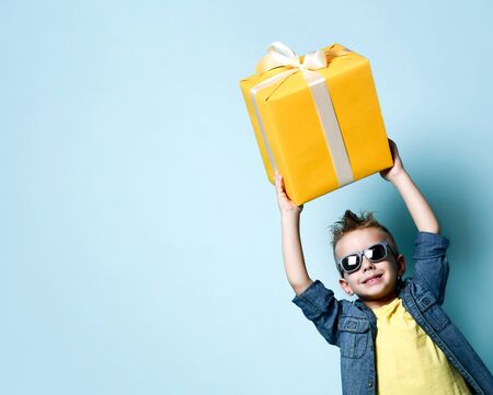 Smiling blond boy in stylish casual clothing and sunglasses holding big yellow present box in raised...