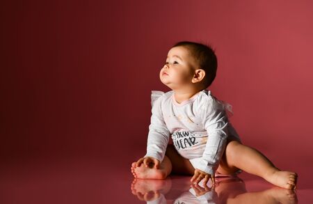 Chubby kid in white bodysuit with inscription, barefoot. She pulling her hands to someone, sitting on floor against pink background. Christmas, New Year, holidays. Advertising for babies. Close upの写真素材