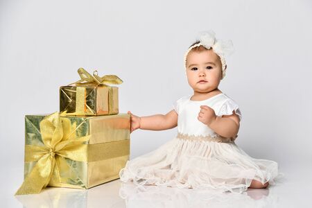 Baby girl in dress and headband, barefoot. She holding golden gift box tied with ribbon, sitting on floor isolated on white. birthday party. Childhood, advertising for babies.の写真素材