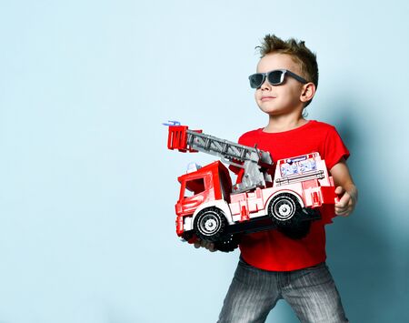 Positive boy in bright stylish casual clothing and sunglasses standing and holding toy fire engine...