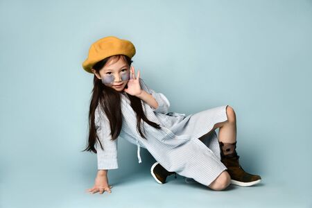 Little brunette asian kid in sunglasses, shirt dress, brown beret, boots. She smiling, posing sitting sideways on floor against blue background. Childhood, fashion, hipster style. Close up, copy spaceの写真素材