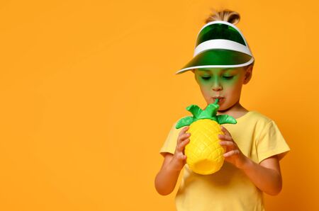 Little smiling cute blond girl in yellow t-shirt and hat drinking fresh healthy fruit juice from...