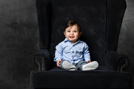 Baby boy in blue shirt, gray pants, white booties. He is about to cry sitting in black armchair on...