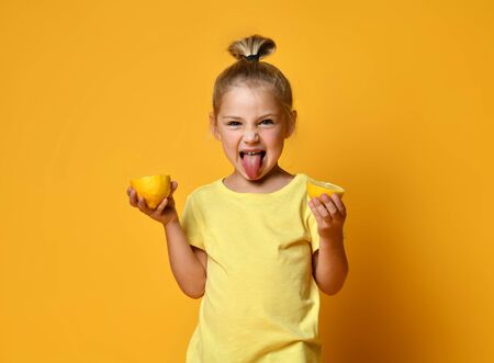 Little smiling cute blond girl in yellow t-shirt holding halves of fresh sour ripe lemon fruit and showing tongue over yellow background. Healthy lifestyle and clean eating conceptの写真素材