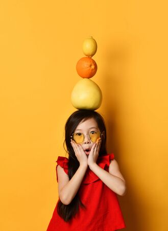 Little asian schoolgirl in sunglasses, red blouse, colored skirt. Amazed, holding pomelo, orange and lemon on her head, posing on orange background. Childhood, fruits, emotions. Close up, copy spaceの写真素材