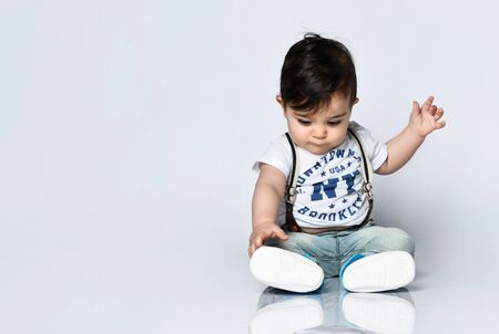 Little child in t-shirt with inscriptions, jeans with suspenders and booties. He is looking wondered, sitting on floor isolated on white. Articles about childhood or advertising for babies. Close upの写真素材