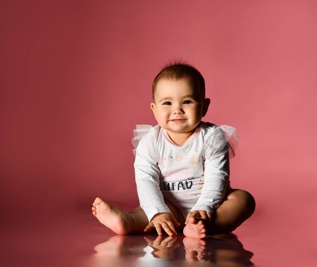 Little child in a smart white bodysuit with inscription, barefoot. She is smiling, sitting on floor against pink background. Christmas, New Year, holidays. Childhood, advertising for babies. Close upの写真素材