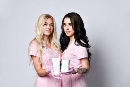 Two ladies in pink medical uniform and latex gloves posing isolated on white. Smiling, holding packages of beauty product with copy space for your text. Cosmetologist, hairdresser. Close upの写真素材