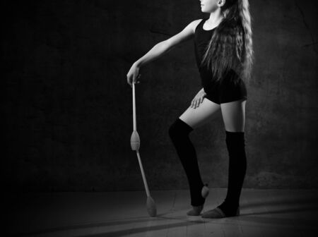 Young gymnast girl in a black body and sport upper arms standing and holding two pink gymnastic maces in hands over gray background. Rhythmic Gymnastics Beauty Conceptの写真素材