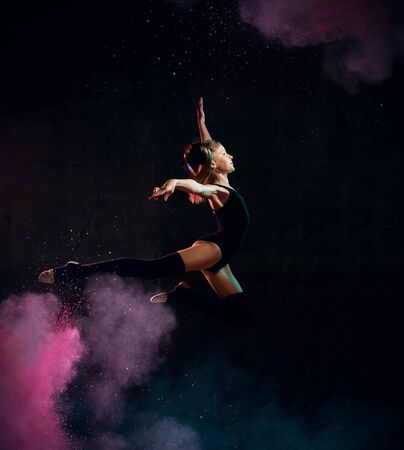 Young girl gymnast in black sport body and special uppers jumping in gymnastic pose over dark background. Rhytmhic gimnastics beauty cocneptの写真素材