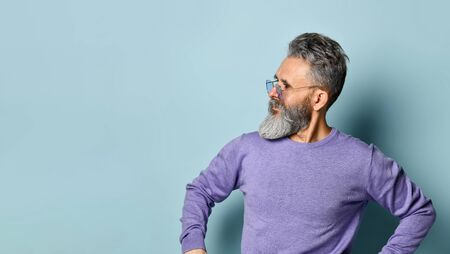 Gray-haired, bearded, elderly man in sunglasses and purple sweater. Hands on hips, looking aside...