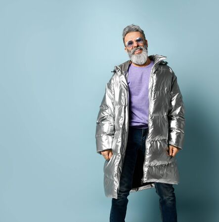 Gray-haired, bearded, mature male in purple pullover and sunglasses, silver colored down jacket, jeans. He is smiling while posing on blue studio background. Fashion, style. Close up, copy spaceの写真素材