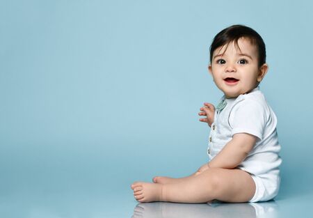 Little kid in white bodysuit as vest with bow-tie, barefoot. He is smiling, sitting on floor against...