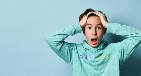 Young teen boy in blue hoodie standing and feeling shocked over pastel blue wall background. Stylish casual clothing for teenagers and emotions diversity conceptの写真素材