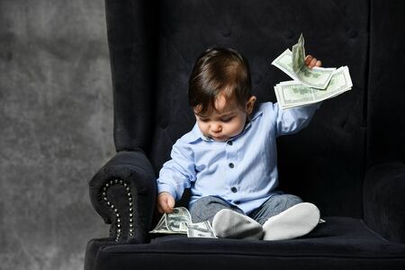 Little baby in blue shirt, gray pants, white booties. He holding some dollar notes, sitting in black armchair on gray background. Articles about childhood or advertisement for toddlers. Close upの写真素材