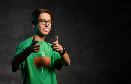 Young smiling teen boy in green t-shirt, jeans and glasses standing and showing good thumbs sign over dark background. Stylish casual clothing for teenagers, expressing emotions conceptの写真素材