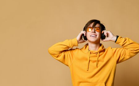Young positive teen boy in yellow hoodie, sunglasses and headphones standing and smiling over yellow wall background. Stylish casual clothing for teenagers and music lover conceptの写真素材