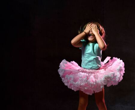 Little female in blue t-shirt and poofy skirt. She is laughing, closed her eyes with palms, posing...
