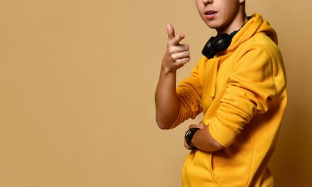 Cropped photo of teen guy in yellow hoodie and headphones standing, smiling and looking at camera...