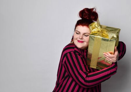 Happy ginger woman in black and purple striped dress, crown and earrings. Holding golden gift box tied with ribbon and bow, looking joyful, posing isolated on white. Close up, copy spaceの写真素材