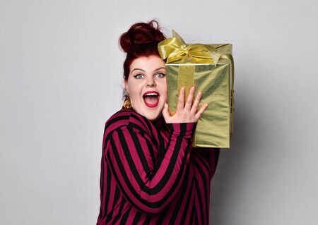 Happy ginger woman in black and purple striped dress, crown and earrings. Holding holds a golden gift box tied to a ribbon and bows in anticipation of surprise.の写真素材